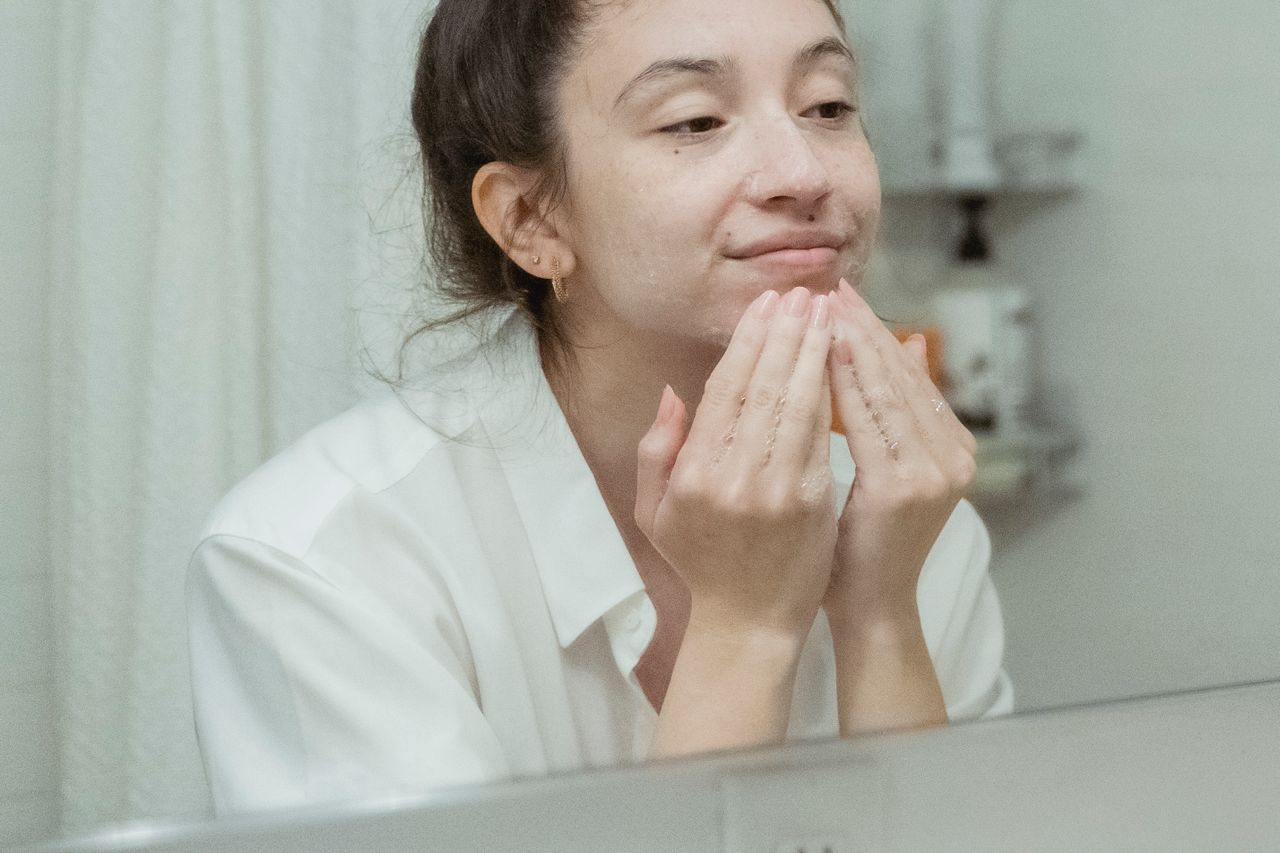 Young woman washing her face with foamy cleanser, reflected in a bathroom mirror, skincare routine